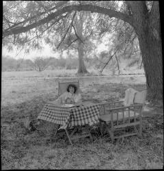 1998.97.571 Jean Buxton sitting at a picnic table in the shade of a large tree at Gogrial, Southern Sudan